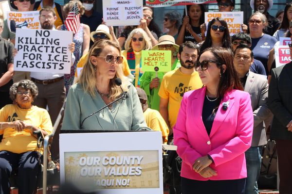 A woman in a green jacket and a woman in a pink jacket stand at a podium in front of a crowd with signs