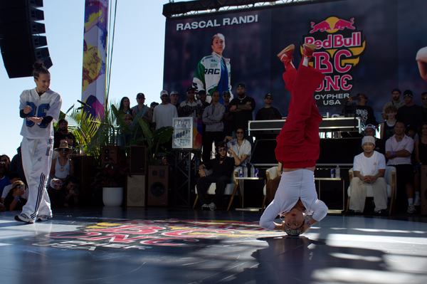 A breaker spins on her head as her opponent watches off to the side miming taking notes. 