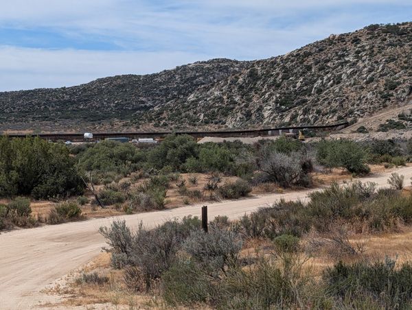 A rocky mountain with a rust brown wall running into it and construction equipment in front, surrounded by scrubby desert