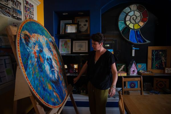 A woman with short brown hair and loose fitting black and green clothing stands in front of a blue and orange mosaic of a bear