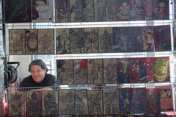 Man smiles surrounded by hand-drawn old school traditional character art which is hung up on his booth in front of him.
