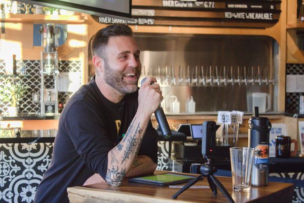 Man smiles while holding mic and leaning on table in front of a bar counter.