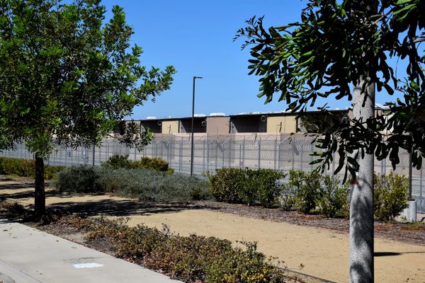 A prison building with a tall fence and razor wire with trees and a dirt path in front