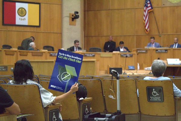 An attendee member facing city committee members holds up a sign that says “Say No to Mass Surveillance, Get the Flock Out.” 