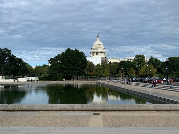 Picture of a capitol building with a dome on top and with trees and a pool of water in front under a cloudy sky