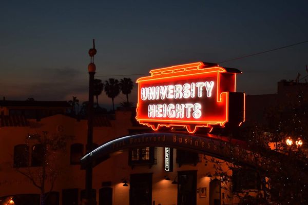 Picture of a sign that says "University Heights" with buildings, palm trees and a sunset behind it.
