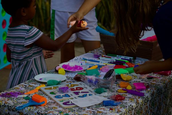 A child passes a toy to a person while someone plays with clay at a table covered in craft supplies.