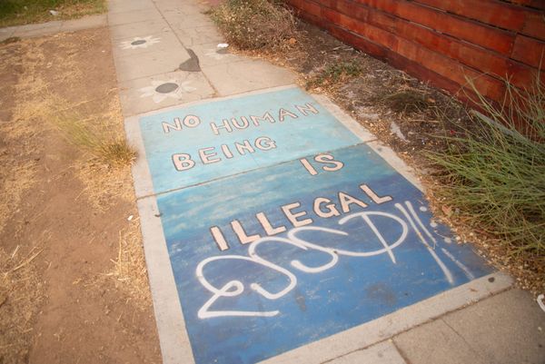 A rectangle of shaded blues on a sidewalk says No Human Being is Illegal
