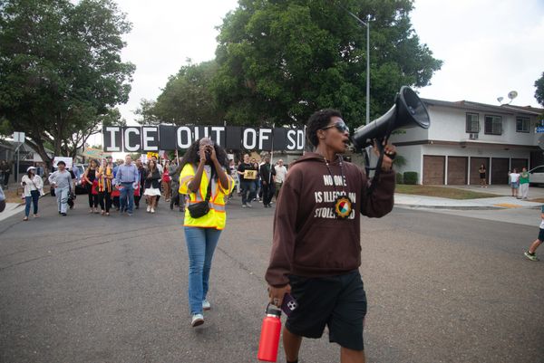 A person holding a megaphone and wearing a brown hoodie leads a crowd carrying signs that say ICE out of San Diego