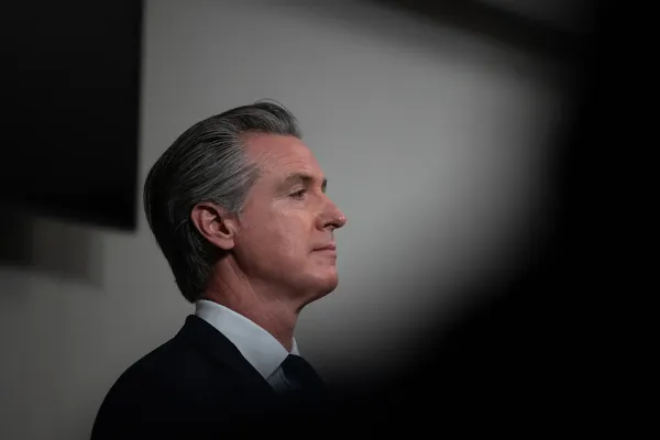 A man with gray hair in a suit stands in front of a white wall