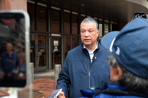 A man speaks outside in front of a building with some facing away from the camera to his right and a cellphone recording on the left