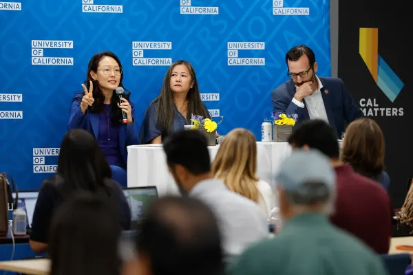 A person holding a microphone in front of a crowd with two other people sitting at a table with them
