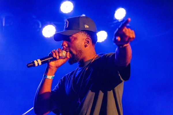 A man in a black shirt and black hat holds a microphone in blue lighting