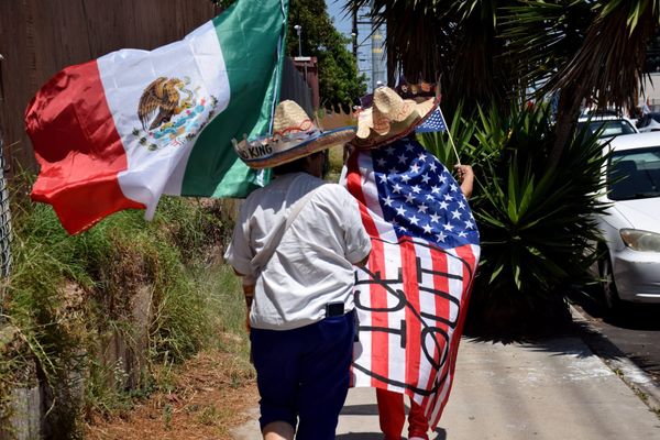 A woman with the American flag as a cape and a woman holding the Mexican flag walk away from the camera with sombreros that say No Kings