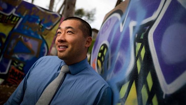 A man in a blue shirt and gray tie stands in front of a purple and yellow graffiti piece