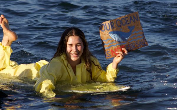 A person with long brown hair wearing a yellow hazmat suit lies on a surf board holding a sign that says our ocean is not for sale