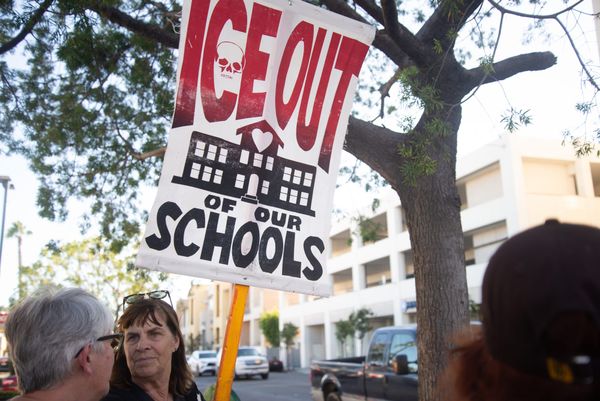 A person holds a red, white and black sign that says ICE Out of Our Schools standing in front of a tree and street