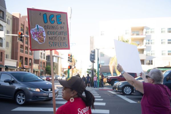 A person in a red shirt and brown hat holds a sign that says "ICE belongs in iced coffee not our streets"