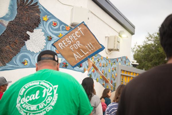 Several people, including one in a green shirt, walk away from the camera, on person holding a sign that says Respect For All. 