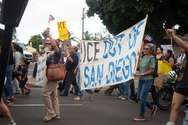 People hold a sign that says in blue and black text "ICE out of San Diego" as they march along a road