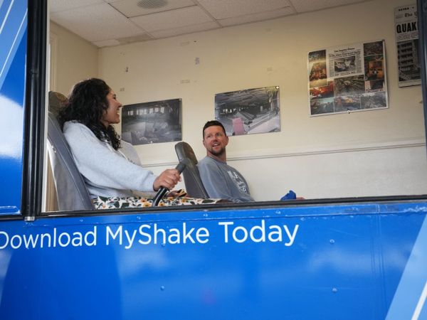 Picture of a simulator device with blue walls with the words "Download MyShake Today" on the outside and white walls inside with a window showing two people sitting in chairs inside