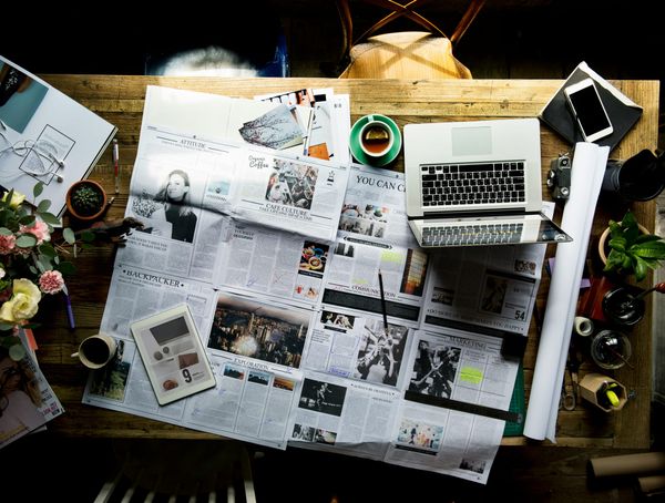 A newspaper chaotically laid out on a desk with coffee, tablet, computer, phone, etc.