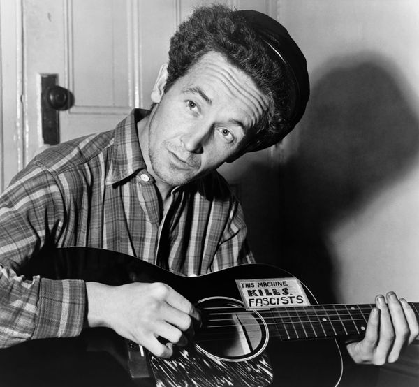 Woody Guthrie playing his guitar with the label "this machine kills fascists" on it.