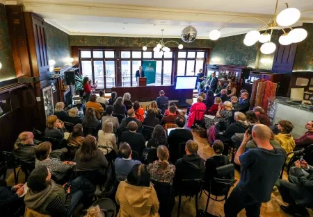 Dans café en boiseries et dorures, le ministre est debout à un pupitre, devant une foule assise.