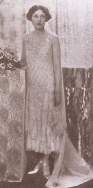 Alt : Barbara Cartland, en pied, du temps de sa jeunesse. Elle porte une robe blanche presque transparente, dont le haut est orné de losanges dorés, et le bas d’arabesques. Une traîne diaphane tombe à ses pieds, chaussés d’escarpins.