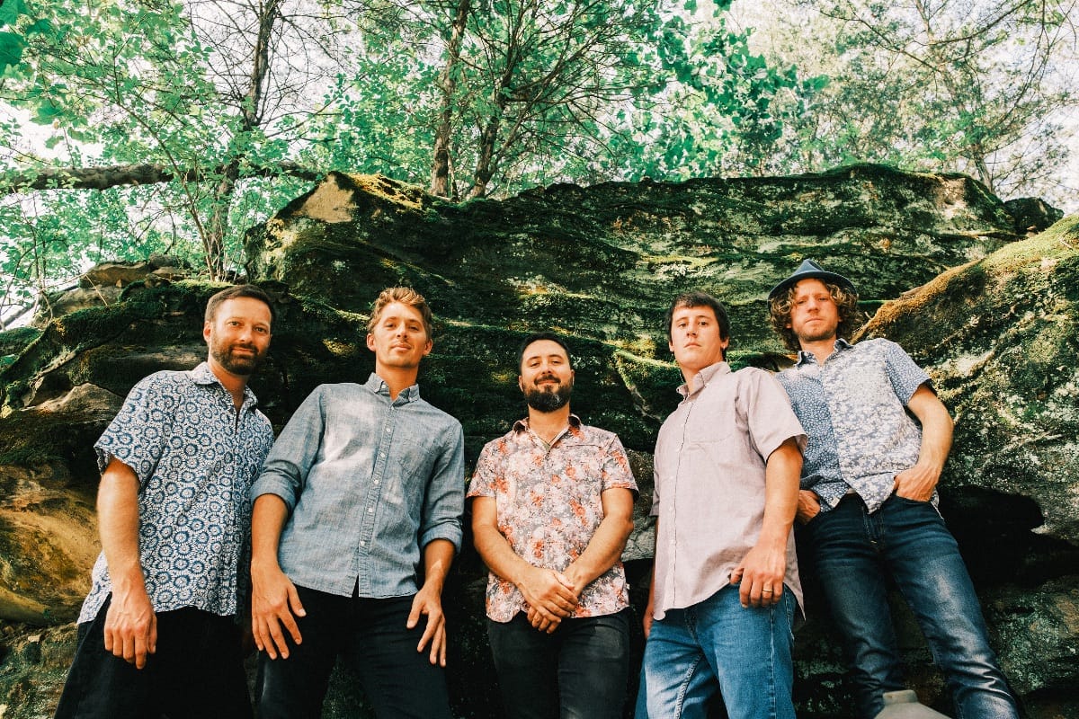 The five members of Fireside Collective stand in a forest by a rock face on a sunny day