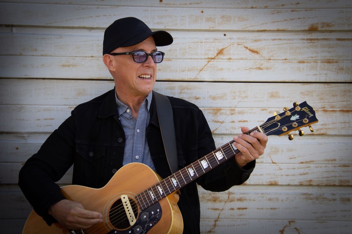 Freedy Johnston against a wood wall holding an acoustic guitar
