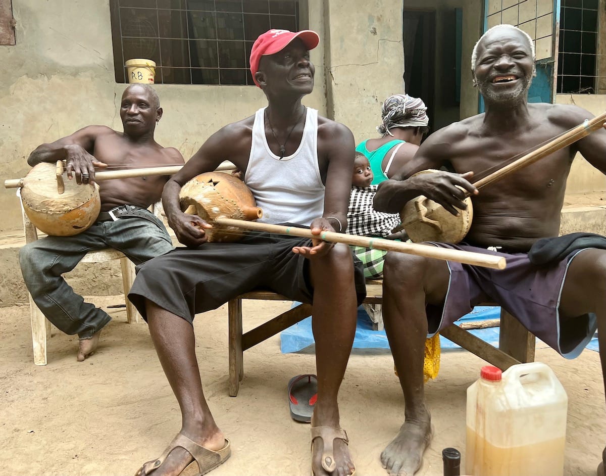 Three men smile and play ekonting, a banjo-like instrument held on their laps.