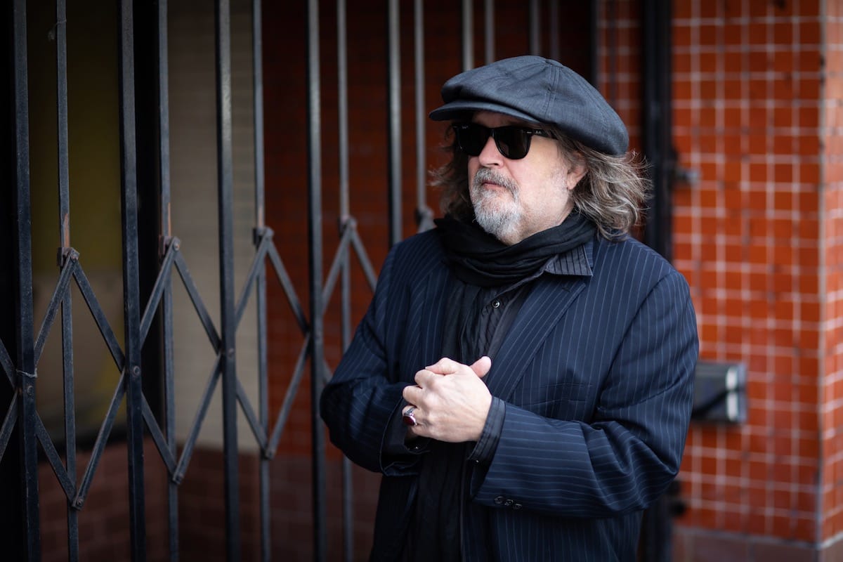 Peter Case in winter clothes standing against a brick wall and black metal grate