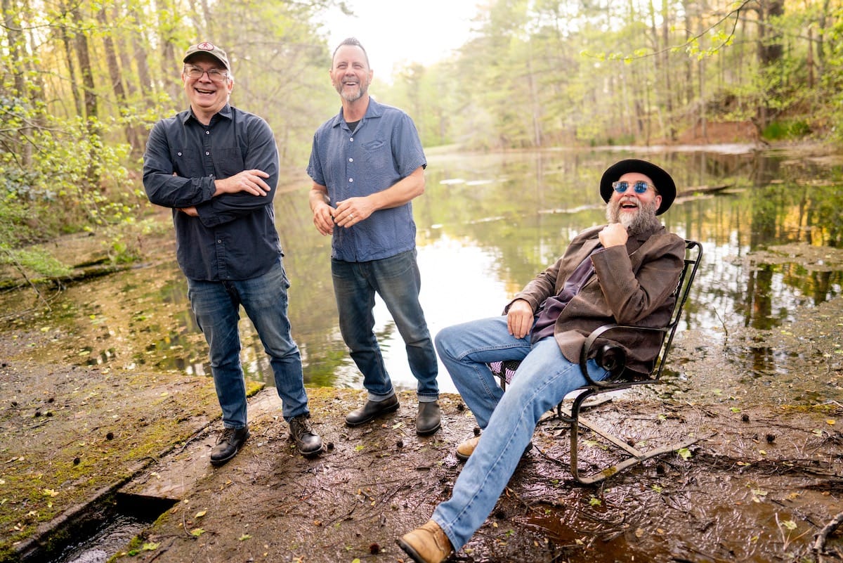 The three members of Chickasaw Mudd Puppies standing or seating on mud next to a pond