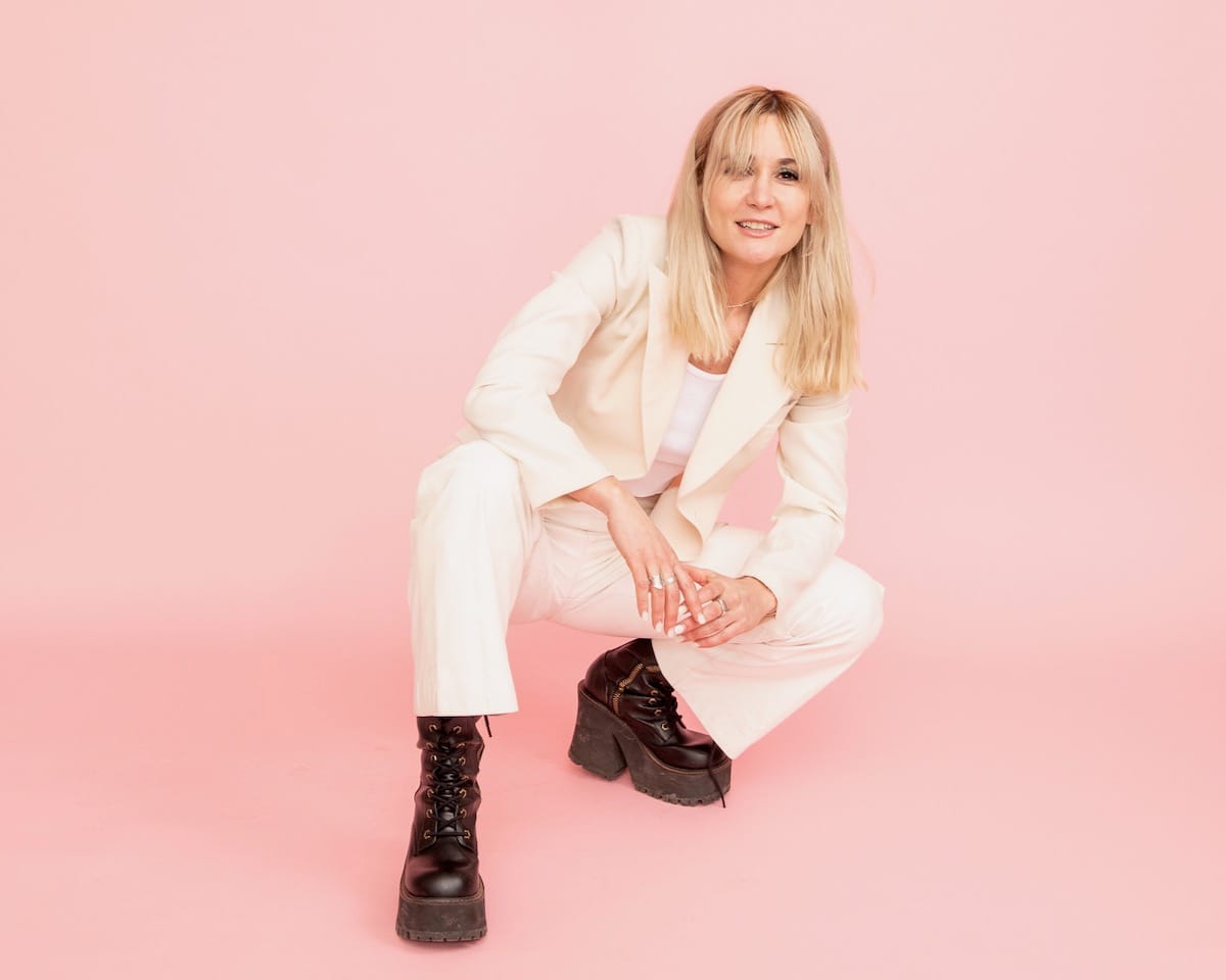 Esther Rose in white suit and black boots against a pink backdrop