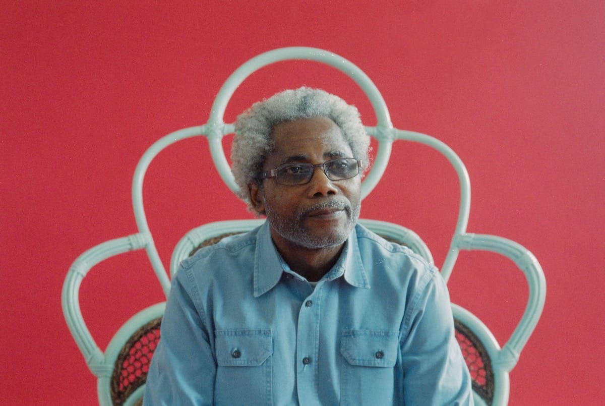 Peter One sits in a cane chair against a red backdrop