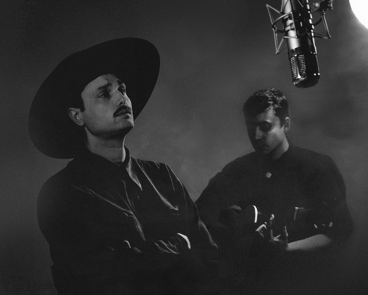Black and white portrait of two men, one singing into a dangling microphone and one playing guitar