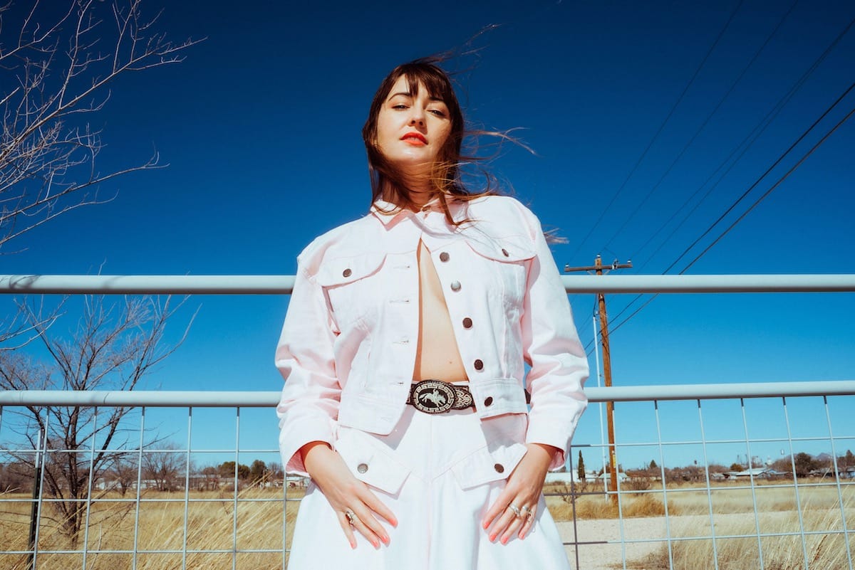 Jess Williamson in white outfit standing against a gray railing and a blue sky