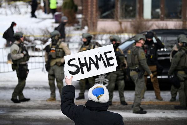 An onlooker holds a sign that reads "Shame" as members of law enforcement work the scene following a suspected shooting by an ICE agent during federal law enforcement operations.