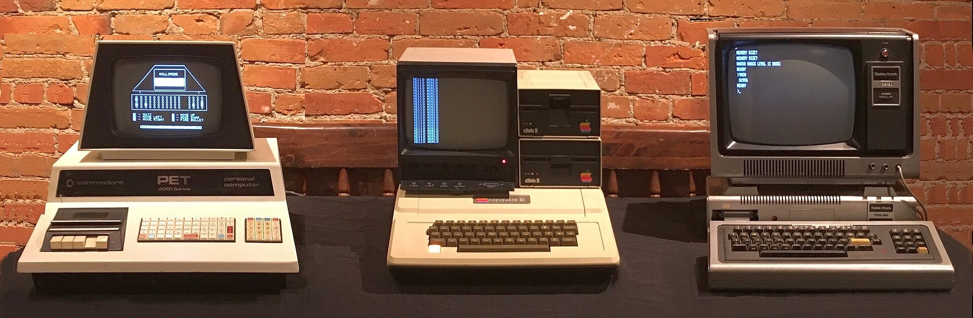 A Commodore PET, Apple 2, and TRS-80 Model 1 are neatly arranged side-by-side, forward facing, on a black table.