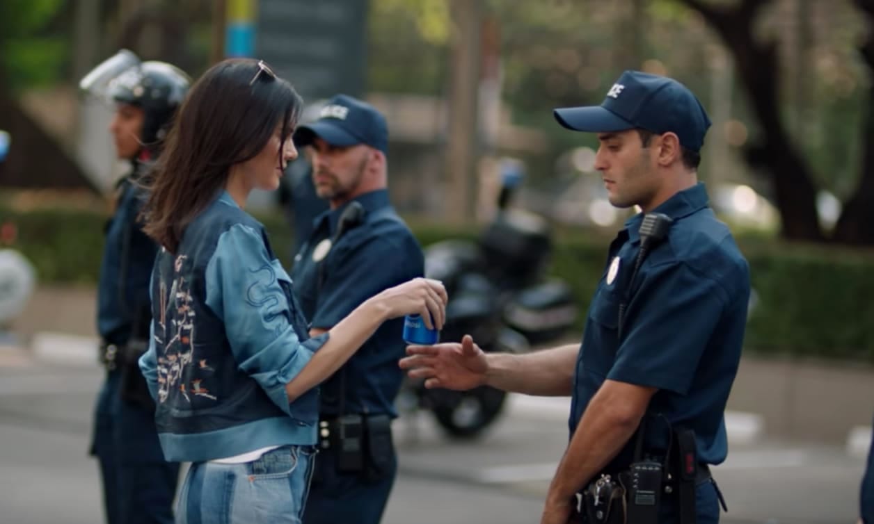 Still frame from the infamous Kylie Jenner Pepsi commercial. Kylie, neatly coiffed and dressed in ever-so-slightly hippie-coded denim, is handing a Pepsi to a cop opposing a protest.