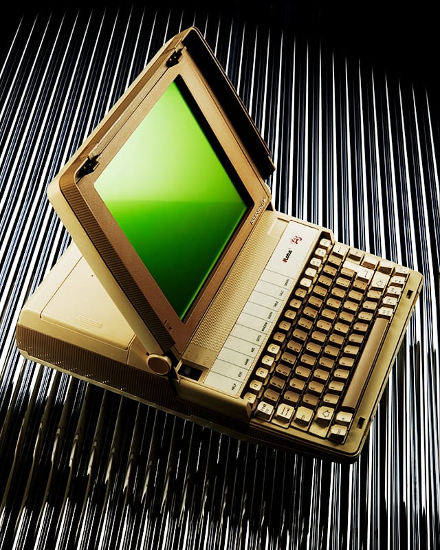 Photo of a Data General/One laptop, shown at an angle against a reflective striped backdrop. The machine has a beige case, a wide green monochrome display, and a full keyboard with chunky 1980s-style keys in stepped rows, thanks to the thickness of the unit. The lid is partially open, highlighting its early portable-computer design.