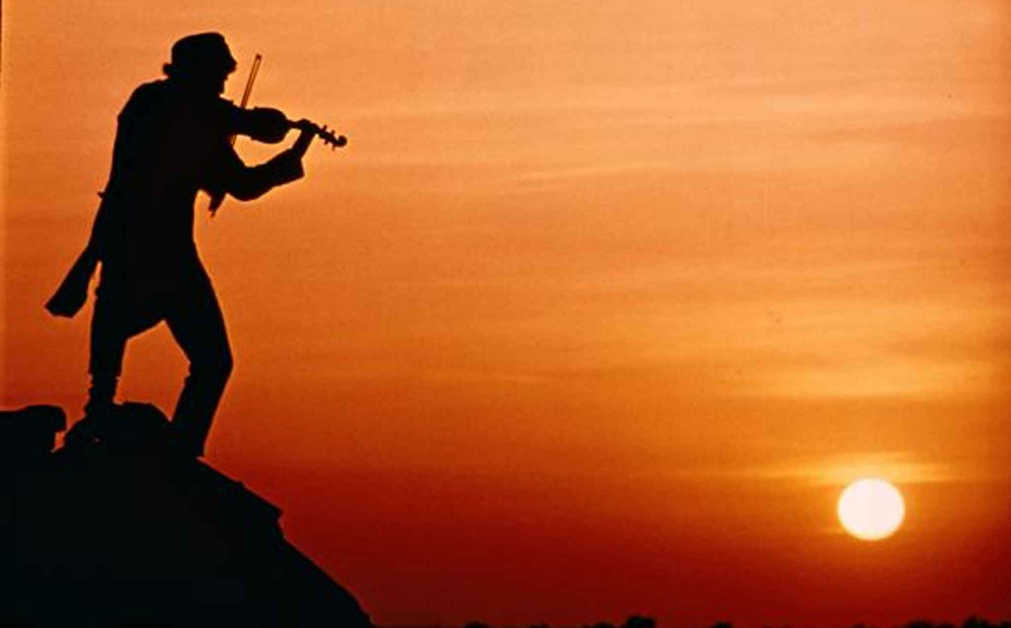 Still frame from the movie Fiddle on the Roof. A silhouette of a man playing a fiddle on a rooftop against a deep orange sunset, the sun sets far off to the right.