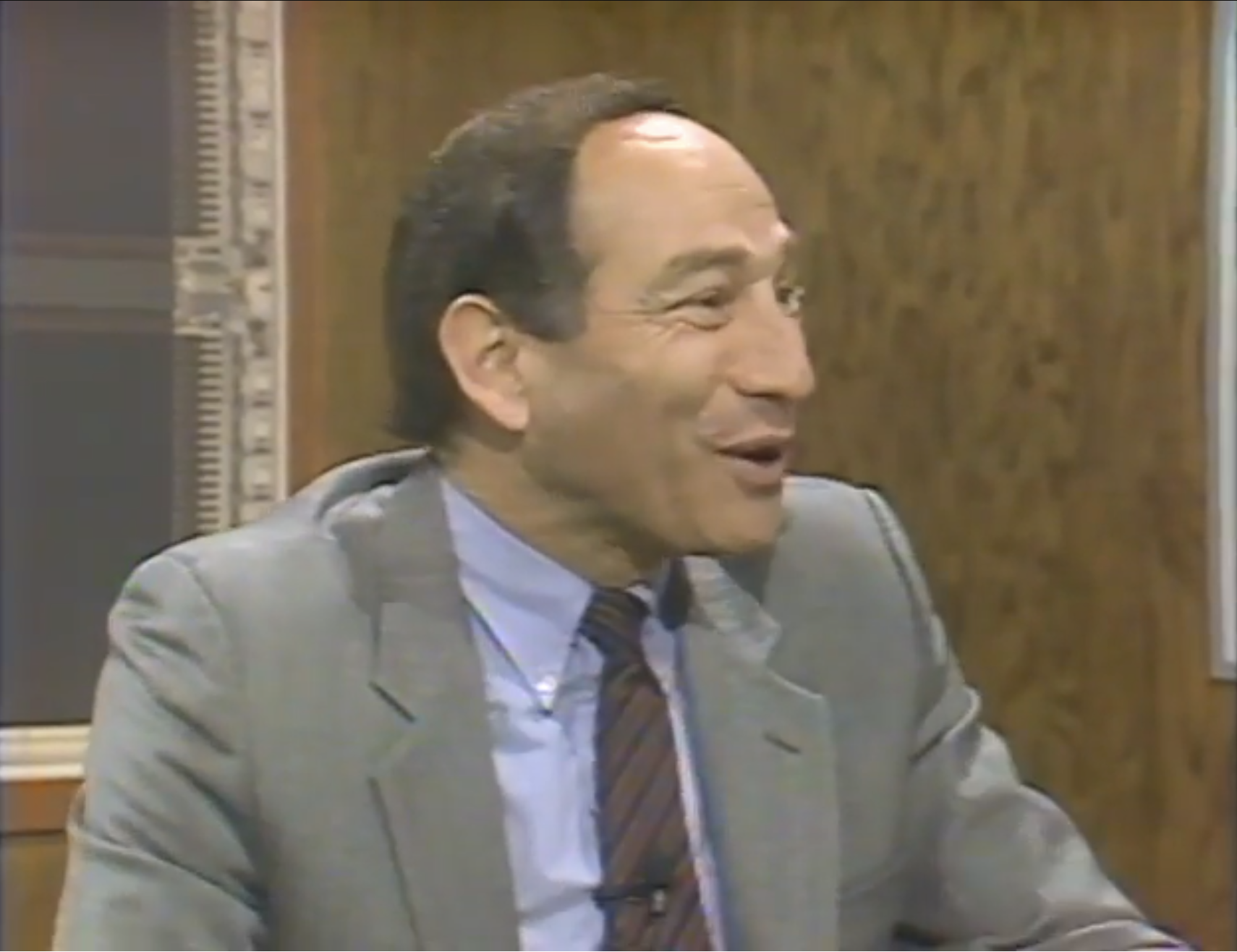 A television host in a gray suit smiles broadly mid-conversation, seated at a desk on a wood-paneled studio set, clearly amused or impressed by what he’s watching or hearing off-camera.
