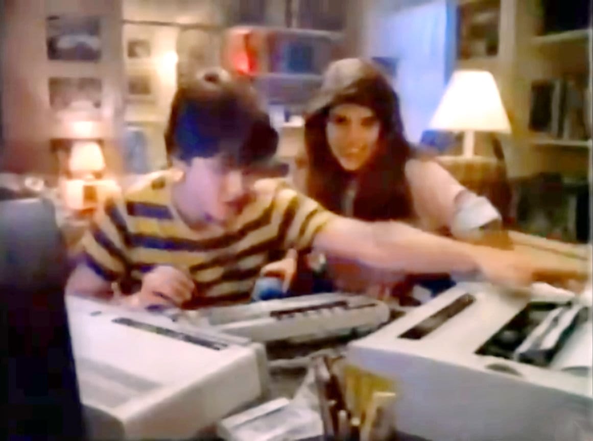 A 1980s television commercial scene showing a teenaged young man using a home computer at a desk while a young woman looks on enthusiastically. A large external printer dominates the foreground, actively printing. She's impressed with his computing skills!