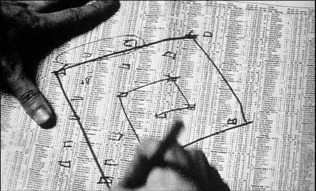A black-and-white film still showing a close-up of a printed numerical table, similar to stock listings or reference data, spread across a page. Two hands enter the frame as one hand draws angular lines and arrows directly over the numbers, forming a rough geometric shape that links scattered points in the data. The image suggests an obsessive attempt to impose structure and meaning onto overwhelming numerical information, visually echoing themes of pattern-seeking and emergent order.