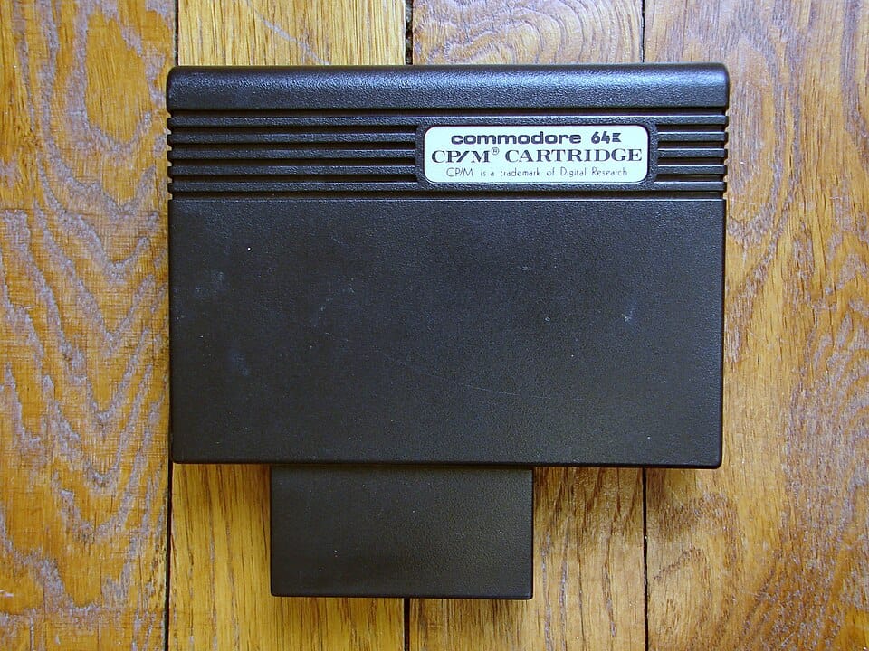 A non-descript black rectangle, a C64 cartridge adorned with a minimal sticker reading, "Commodore 64 CP/M Cartridge" The cart sits on a wooden table. 