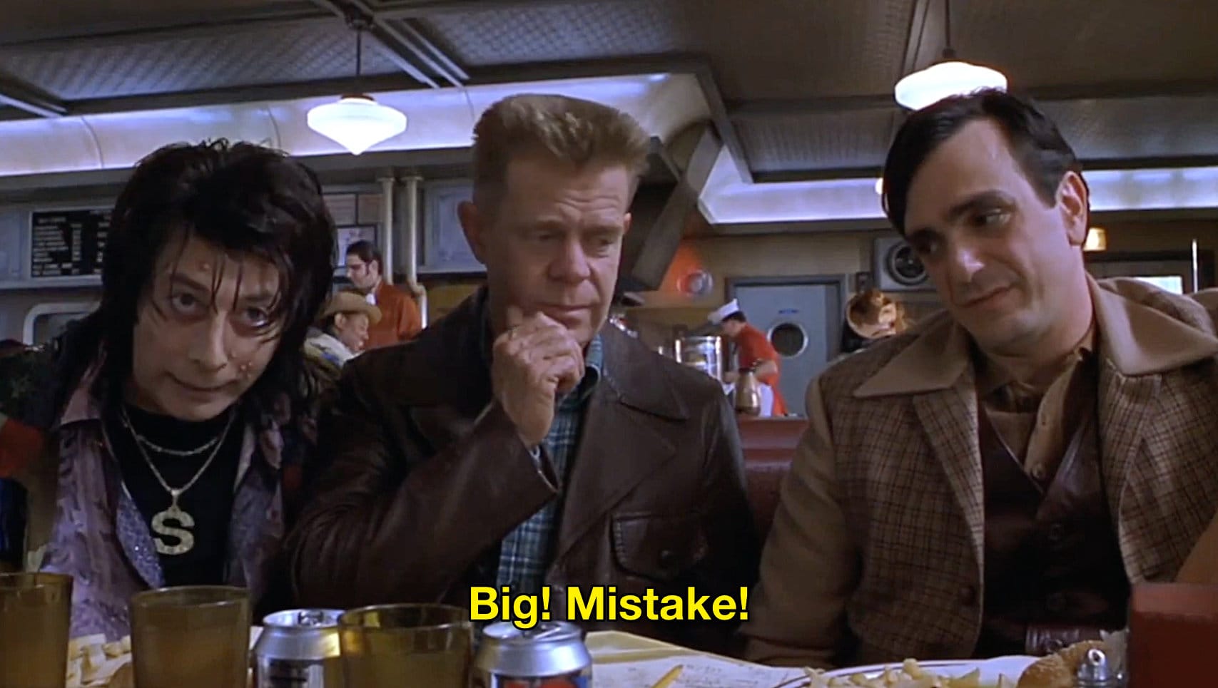 Frame grab from the movie Mystery Men. A retro 50s style diner scene, three men are squeezed into a booth. Paul Reubens on the far left as The Spleen recounts his origin story, decked out in flamboyant sparkly clothes and a gold "S" on a chain, jaundiced skin, emo haircut. William Macy in the middle, in a leather jacket and plaid shirt, short high top haircut, as The Shoveler turns away from Spleen. On the far right, Hank Azaria as The Blue Raja in a 70s style wide collar brown suit with vest looks annoyed at having to hear the story again.