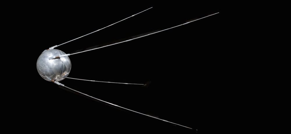 An image of the Sputnik, a silver sphere with four long, thin spikes flared out in a pyramid shape from the ball. It shines against a pure black background.