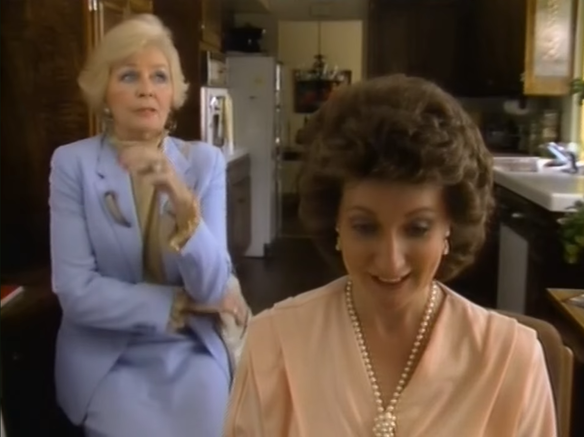 Still from the dBASE video, set in a kitchen. A well-dressed woman wearing a pearl necklace sits in the foreground, smiling as she plugs away at dBASE, while an older woman in a light-blue suit sits on a stool behind her, arms crossed, mid-eye-roll.